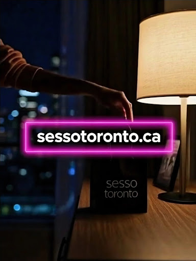 A person unpacking a Sesso Toronto shopping bag on a wooden table next to a lamp at night with a city view in the background.